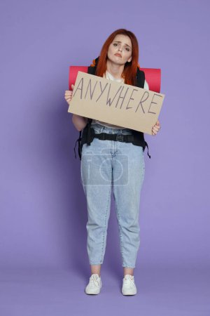 Photo for Tourism and active free time advertisement concept. Portrait of girl traveler. Woman tourist in casual with backpack sad face, holding sign with word anywhere on it. Isolated on purple background. - Royalty Free Image