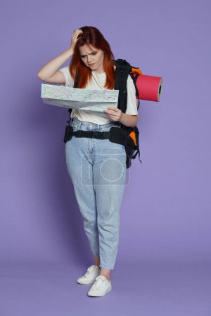 Photo for Tourism and active free time advertisement concept. Portrait of girl traveler. Woman tourist in casual with backpack holding paper map looking confused, holding head. Isolated on purple background. - Royalty Free Image