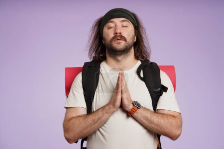 Photo for Tourism and active leisure time advertisement concept. Portrait of male traveler. Man tourist in casual clothing with backpack holding hands in praying position. Isolated on purple background. - Royalty Free Image