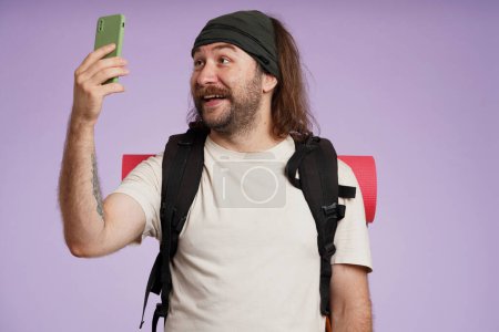 Photo for Tourism and active leisure time advertisement concept. Portrait of male traveler. Man tourist in casual clothing with backpack holding smartphone taking selfie, smiling. Isolated on purple background. - Royalty Free Image