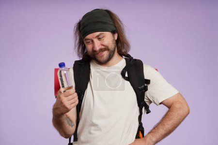 Photo for Tourism and active leisure time advertisement concept. Portrait of male traveler. Man tourist in casual clothing with backpack smiling, holding plastic bottle with water. Isolated on purple background - Royalty Free Image
