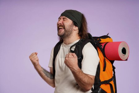 Photo for Tourism and active leisure time advertisement concept. Portrait of male traveler. Man tourist in casual clothing with backpack happy screaming, showing winning gesture. Isolated on purple background. - Royalty Free Image