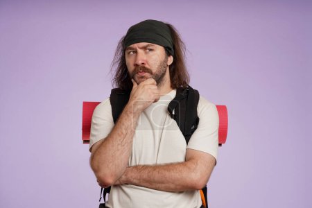 Photo for Tourism and active leisure time advertisement concept. Portrait of male traveler. Man tourist in casual clothing with backpack stands with crossed hands and thinking. Isolated on purple background. - Royalty Free Image