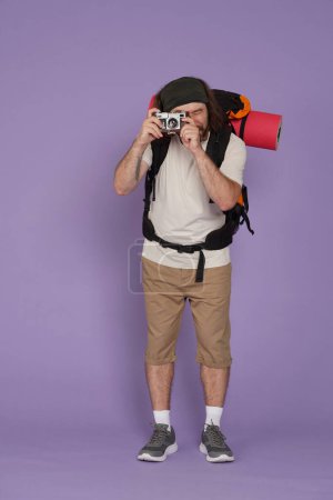 Photo for Tourism and active leisure time advertisement concept. Portrait of male traveler. Man tourist in casual clothing with backpack and old film camera, taking pics. Isolated on purple background. - Royalty Free Image