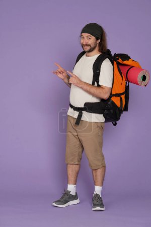 Photo for Tourism and active leisure time advertisement concept. Portrait of male traveler. Man tourist in casual clothing with backpack standing pointing at empty area, smiling. Isolated on purple background. - Royalty Free Image