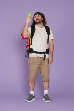Photo for Tourism and active leisure time advertisement concept. Portrait of male traveler. Man tourist in casual clothing with backpack holding smartphone looking for signal. Isolated on purple background. - Royalty Free Image