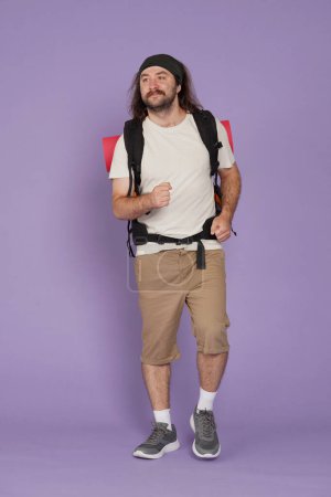 Photo for Tourism and active leisure time advertisement concept. Portrait of male traveler. Man tourist in casual clothing with backpack walks, positive expression, looks around. Isolated on purple background. - Royalty Free Image