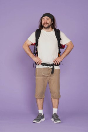 Photo for Tourism and active leisure time advertisement concept. Portrait of male traveler. Man tourist in casual clothing with backpack looking at the camera smiling expression. Isolated on purple background. - Royalty Free Image
