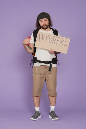 Photo for Tourism and active leisure time advertisement concept. Portrait of male traveler. Man tourist in casual with backpack holding paper sign with word anywhere written on it. Isolated on purple background - Royalty Free Image