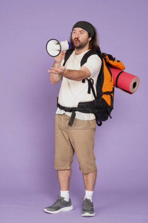 Photo for Tourism and active leisure time advertisement concept. Portrait of male traveler. Man tourist in casual clothing with backpack talking in megaphone with serious face. Isolated on purple background. - Royalty Free Image
