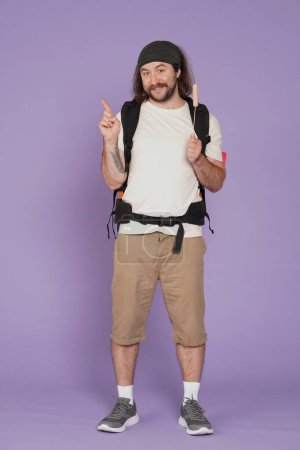 Photo for Tourism and active leisure time advertisement concept. Portrait of male traveler. Man tourist in casual with backpack holds sausage on stick and pointing at empty area. Isolated on purple background. - Royalty Free Image