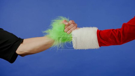 Photo for Greeting gestures of man and Grinchs hairy green hand on isolated blue background. Gift kidnapper cosplay. Christmas and New Year celebration concept. Blue screen, chroma key - Royalty Free Image