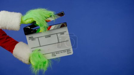 Foto de La mano de pelo verde Grinchs sostiene un aplauso de película sobre un fondo azul aislado. Cosplay secuestrador de regalos. Concepto de celebración de Navidad y Año Nuevo. Pantalla azul, croma key - Imagen libre de derechos