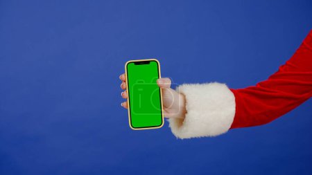 Photo for Female hand in Santa Claus costume with green screen phone on isolated blue background. Template place for your text or image, promotional content. Advertising area, workspace mock up. - Royalty Free Image