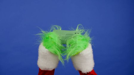 Foto de Grinchs manos de pelo verde juegan un juego en un teléfono inteligente sobre un fondo azul aislado. Cosplay secuestrador de regalos. Concepto de celebración de Navidad y Año Nuevo. Pantalla azul, croma key - Imagen libre de derechos