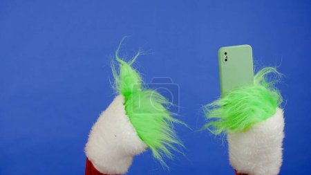Foto de La mano de pelo verde Grinchs sostiene el teléfono inteligente y muestra un gesto hacia arriba pulgares. Cosplay secuestrador de regalos. Concepto de celebración de Navidad y Año Nuevo. Pantalla azul, croma key - Imagen libre de derechos
