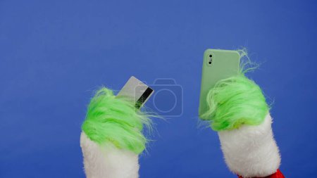 Téléchargez les photos : Grinchs mains aux cheveux verts tenant une carte bancaire et un téléphone intelligent sur fond bleu isolé. Cadeau kidnappeur cosplay. Noel et Nouvel An concept de célébration. Écran bleu, clé chromatique - en image libre de droit