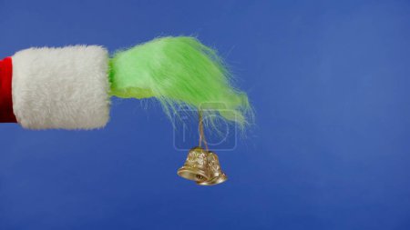 Foto de La mano de pelo verde Grinchs sostiene campanas doradas sobre un fondo azul aislado. Cosplay secuestrador de regalos. Concepto de celebración de Navidad y Año Nuevo. Pantalla azul, croma key. - Imagen libre de derechos