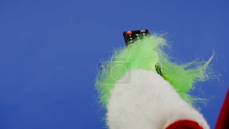 Photo for The Grinchs green haired hand changes channels on a TV using a remote on an isolated blue background. Gift kidnapper cosplay. Christmas and New Year celebration concept. Blue screen, chroma key - Royalty Free Image