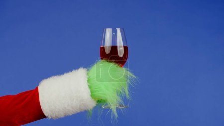 Foto de La mano de pelo verde Grinchs sostiene una copa de vino tinto sobre un fondo azul aislado. Cosplay secuestrador de regalos. Concepto de celebración de Navidad y Año Nuevo. Pantalla azul, croma key - Imagen libre de derechos
