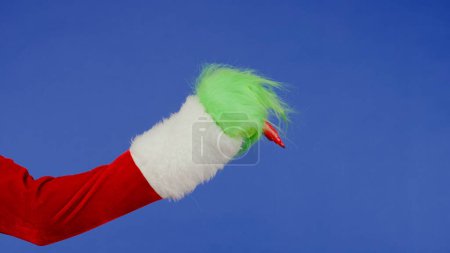 Foto de La mano de pelo verde Grinchs sostiene el marcador rojo sobre un fondo azul aislado. Cosplay secuestrador de regalos. Concepto de celebración de Navidad y Año Nuevo. Pantalla azul, croma key - Imagen libre de derechos
