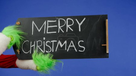 Foto de Las manos de pelo verde Grinchs sostienen un letrero negro que dice Feliz Navidad sobre un fondo azul aislado. Cosplay secuestrador de regalos. Concepto de celebración de Año Nuevo. Pantalla azul, croma key. - Imagen libre de derechos