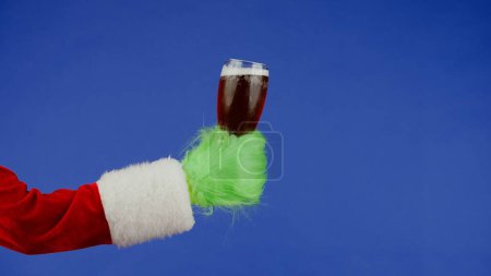 Foto de Grinchs mano de pelo verde sostiene un vaso de cerveza oscura sobre un fondo azul aislado. Cosplay secuestrador de regalos. Concepto de celebración de Navidad y Año Nuevo. Pantalla azul, croma key - Imagen libre de derechos