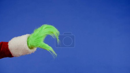 Photo for The Grinchs green haired hand shows a half heart sign with his fingers on an isolated blue background. Gift snatcher cosplay. Christmas and New Year celebration concept. Blue screen, chroma ke - Royalty Free Image
