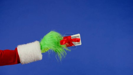 Photo for Green haired Grinch hand holds a wad of money wrapped in a red bow on an isolated background. A place for your advertisement. Christmas and new year celebration concept. Blue screen, chroma key - Royalty Free Image