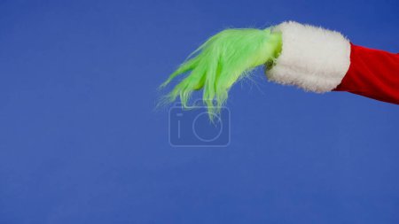 Photo for The Grinchs green hairy hand seems to be holding something against an isolated blue background. Gift snatcher cosplay. Christmas and New Year celebration concept. Blue screen, chroma key - Royalty Free Image