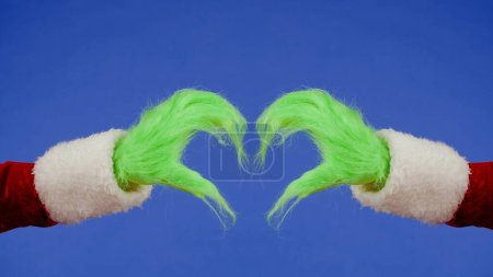 Foto de Manos peludas verdes mostrando el signo del corazón con los dedos sobre fondo azul aislado. Cosplay secuestrador de regalos. Navidad y año nuevo concepto de celebración. Pantalla azul, croma key - Imagen libre de derechos