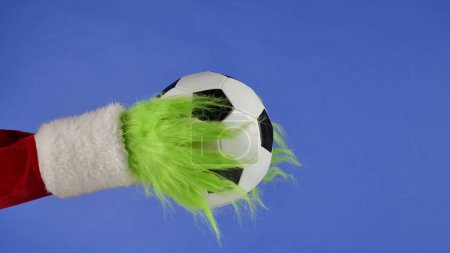 Téléchargez les photos : Grinches vertes mains poilues tenant un ballon de football sur fond bleu isolé. Un cosplay de voleur de cadeaux. Noël et le concept de célébration du Nouvel An. Écran bleu, clé chromatique - en image libre de droit
