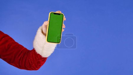 Photo for Female hand in Santa Claus costume with green screen phone on isolated blue background. Template place for your text or image, promotional content. Advertising area, workspace mock up. - Royalty Free Image