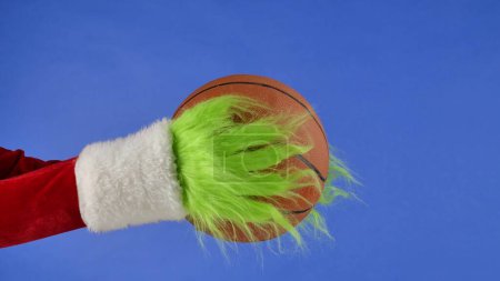 Téléchargez les photos : Grinches vertes mains poilues tenant un ballon de basket sur fond bleu isolé. Un cosplay de voleur de cadeaux. Noël et le concept de célébration du Nouvel An. Écran bleu, clé chromatique - en image libre de droit