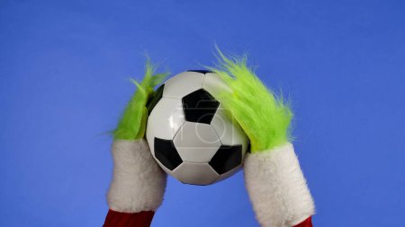 Foto de Grinchs manos peludas verdes sosteniendo una pelota de fútbol sobre fondo azul aislado. Cosplay secuestrador de regalos. Navidad y año nuevo concepto de celebración. Pantalla azul, croma key - Imagen libre de derechos