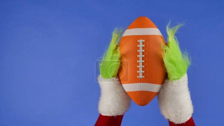 Téléchargez les photos : Grinches vertes poilues mains tenant un soccer américain sur fond bleu isolé. Un cosplay de voleur de cadeaux. Noël et le concept de célébration du Nouvel An. Écran bleu, clé chromatique - en image libre de droit