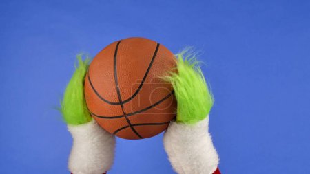 Téléchargez les photos : Grinches vertes mains poilues tenant un ballon de basket sur fond bleu isolé. Un cosplay de voleur de cadeaux. Noël et le concept de célébration du Nouvel An. Écran bleu, clé chromatique - en image libre de droit