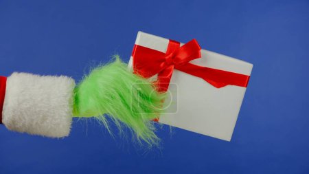 Foto de La mano de pelo verde Grinchs sostiene una caja de regalo blanca con un lazo rojo sobre un fondo azul aislado. Cosplay secuestrador de regalos. Concepto de celebración de Navidad y Año Nuevo. Pantalla azul, croma key. - Imagen libre de derechos