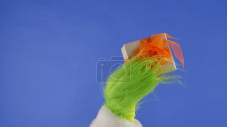 Téléchargez les photos : La main aux cheveux verts Grinchs tient une boîte cadeau blanche avec un noeud orange sur un fond bleu isolé. Cadeau kidnappeur cosplay. Noel et Nouvel An concept de célébration. Écran bleu, clé chromatique. - en image libre de droit
