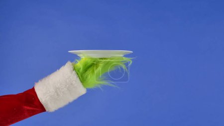 Téléchargez les photos : La main aux cheveux verts Grinchs montre une plaque blanche sur un fond bleu isolé. Un cosplay de voleur de cadeaux. Noël et le concept de célébration du Nouvel An. Écran bleu, clé chromatique - en image libre de droit