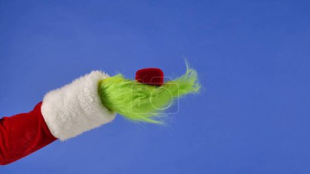 Foto de Manos peludas verdes del Grinch sosteniendo un joyero de terciopelo rojo sobre un fondo azul aislado. Cosplay ladrón de regalos. Concepto de celebración de Navidad y Año Nuevo. Pantalla azul, tecla de cromo - Imagen libre de derechos