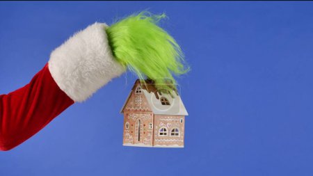 Foto de La mano de pelo verde Grinchs sostiene una casa decorativa nevada sobre un fondo azul aislado. Robador de regalos Cosplay. Concepto de celebración de Navidad y Año Nuevo. Pantalla azul, croma key - Imagen libre de derechos