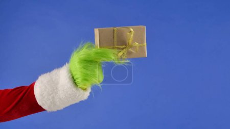 Photo for The Grinchs green haired hand holds a white gift box with a gold bow on an isolated blue background. Gift kidnapper cosplay. Christmas and New Year celebration concept. Blue screen, chroma key. - Royalty Free Image