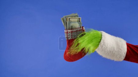 Photo for The Grinchs green haired hand hold a Christmas sock with dollar bills on a blue isolated background. Gift Snatcher Cosplay. Christmas and New Year celebration concept. Blue screen, chroma key - Royalty Free Image