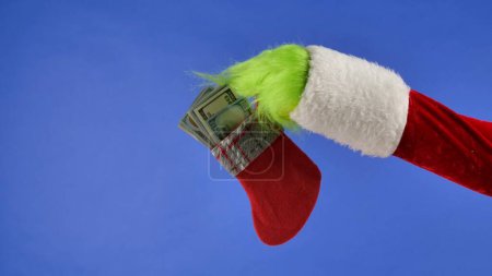 Téléchargez les photos : La main aux cheveux verts Grinchs tient une chaussette de Noël avec des billets d'un dollar sur un fond bleu isolé. Cadeau Snatcher Cosplay. Noel et Nouvel An concept de célébration. Écran bleu, clé chromatique - en image libre de droit