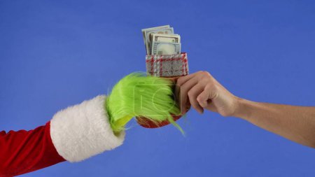 Foto de La mano de pelo verde Grinchs sostiene un calcetín de Navidad con billetes de dólar y una mano de hombre se lo lleva. Robador de regalos Cosplay. Concepto de celebración de Navidad y Año Nuevo. Pantalla azul, croma key - Imagen libre de derechos