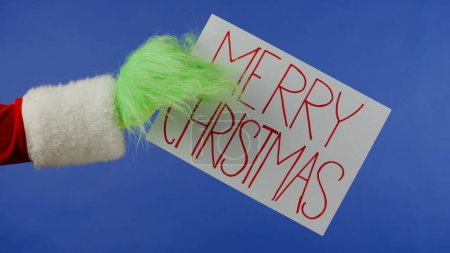 Foto de La mano de pelo verde Grinchs sostiene un cartel blanco que dice Feliz Navidad sobre un fondo azul aislado. Cosplay secuestrador de regalos. Concepto de celebración de Año Nuevo. Pantalla azul, croma key. - Imagen libre de derechos