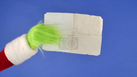 Photo for The Grinchs green haired hand holds a blank vintage kraft paper on a blue isolated background. Gift Snatcher Cosplay. Christmas and New Year celebration concept. Blue screen, chroma key - Royalty Free Image