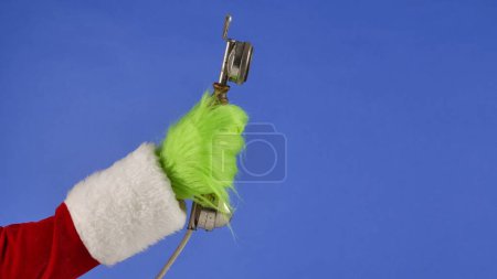 Foto de La mano de pelo verde Grinchs sostiene un receptor de teléfono antiguo sobre un fondo azul aislado. Robador de regalos Cosplay. Concepto de celebración de Navidad y Año Nuevo. Pantalla azul, croma key - Imagen libre de derechos