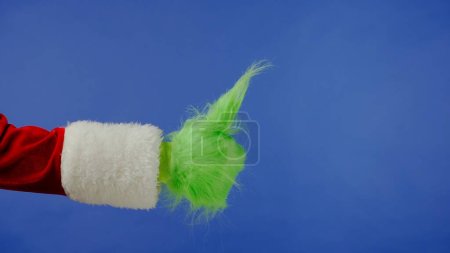 Foto de Mano Grinch de pelo verde muestra pulgares hacia arriba gesto sobre fondo azul aislado. Cosplay secuestrador de regalos. Concepto de celebración de Navidad y Año Nuevo. Pantalla azul, croma key. - Imagen libre de derechos